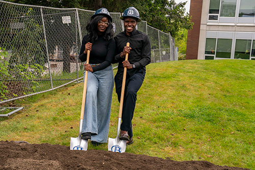 Two people pose with shovels at the dirt mound. 