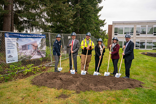 Six people wearing hard hats pose with shovels in dirt with a fence and sign to the left.