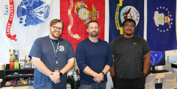 Three people stand in front of military flags.