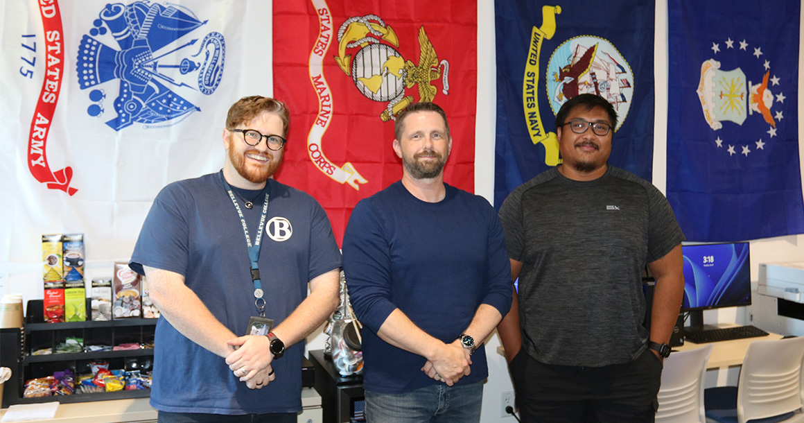Three people stand in front of military flags.