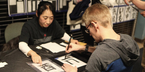 Two people sit at a table while one draws a Japanese character.