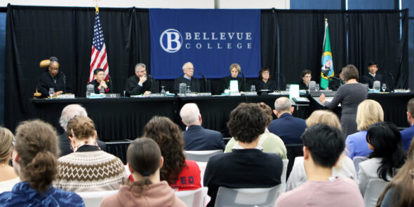 A formal event at Bellevue College featuring a judicial panel seated behind a long table on a stage. The backdrop displays the Bellevue College logo, flanked by the U.S. flag on the left and the Washington State flag on the right. Several judges are seated at the table with nameplates in front of them, and a person stands at a podium addressing the panel. In the foreground, an audience is seated and facing the stage.