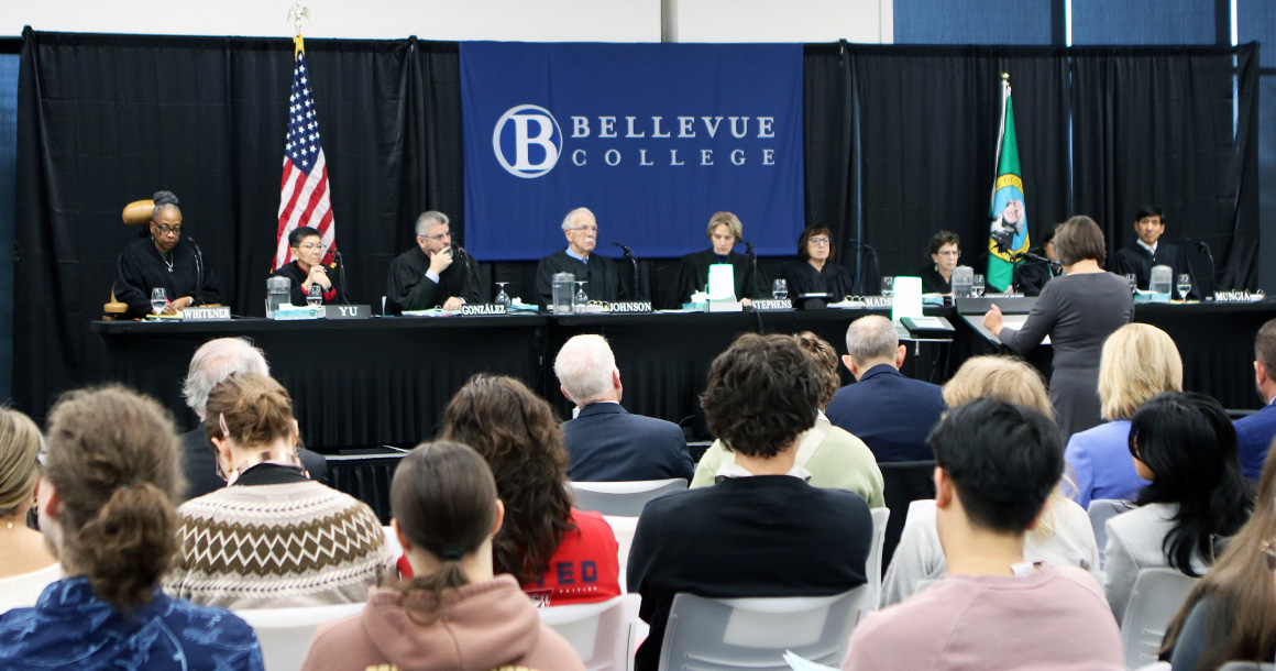A formal event at Bellevue College featuring a judicial panel seated behind a long table on a stage. The backdrop displays the Bellevue College logo, flanked by the U.S. flag on the left and the Washington State flag on the right. Several judges are seated at the table with nameplates in front of them, and a person stands at a podium addressing the panel. In the foreground, an audience is seated and facing the stage.