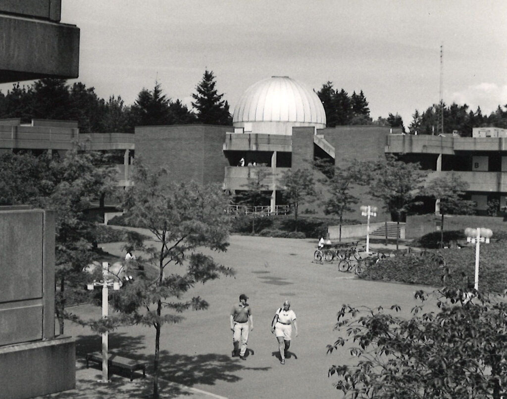 A black and white image of Bellevue College's campus with two people walking and the planetarium in the background. 