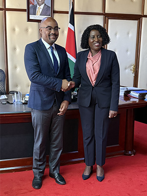 Two people stand in front of a desk, shaking hands and smiling.