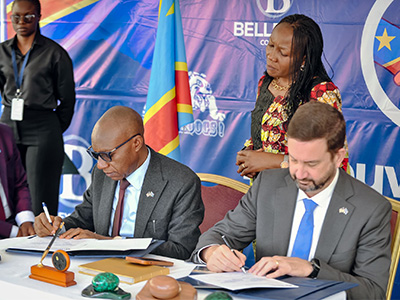 Two men sit at a table, signing papers with a pen, as two people behind them watch.
