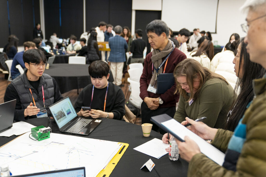 Six people gather around a table with a computer as a person talks.