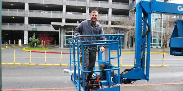 Person stands in the basket of a blue crane in front of the parking garage.