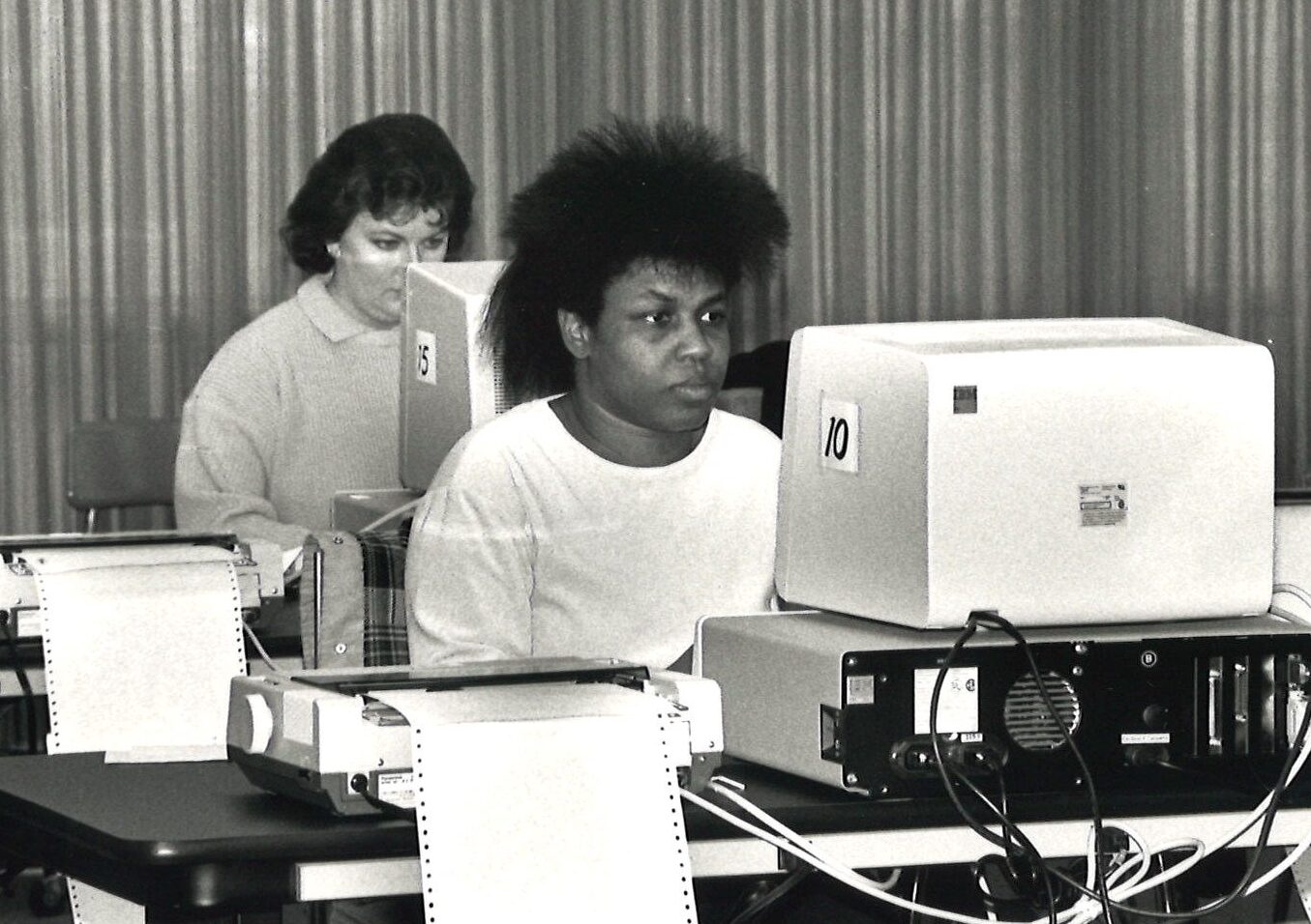 Two people sit at computers with printers and paper next to them.