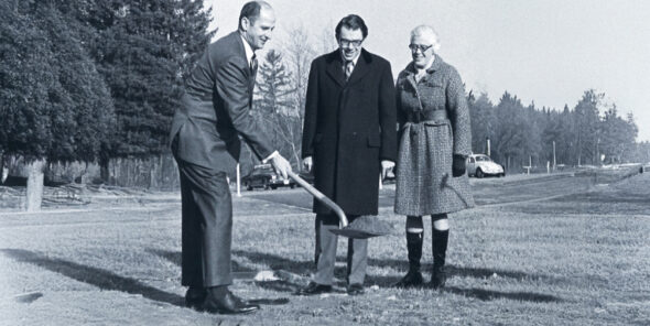 One person holds a shovel while two people look on outside in front of a parking lot.