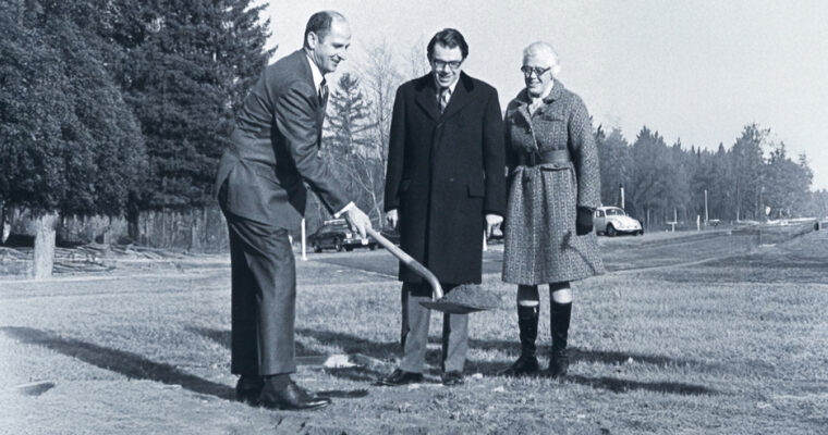 One person holds a shovel while two people look on outside in front of a parking lot.
