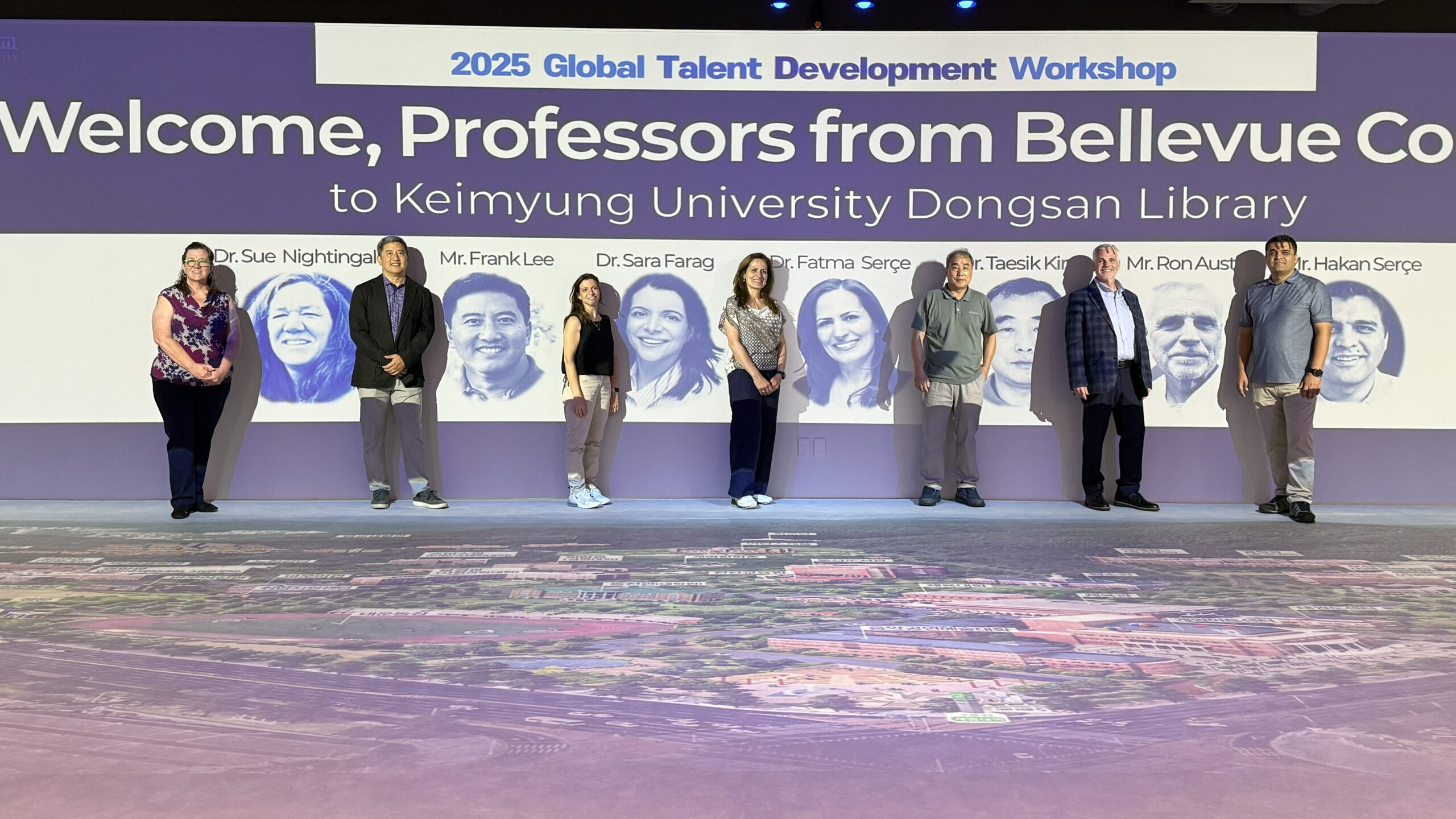 Seven instructors stand next to a wall with designed pictures of their faces, a welcome from Keimyung University.