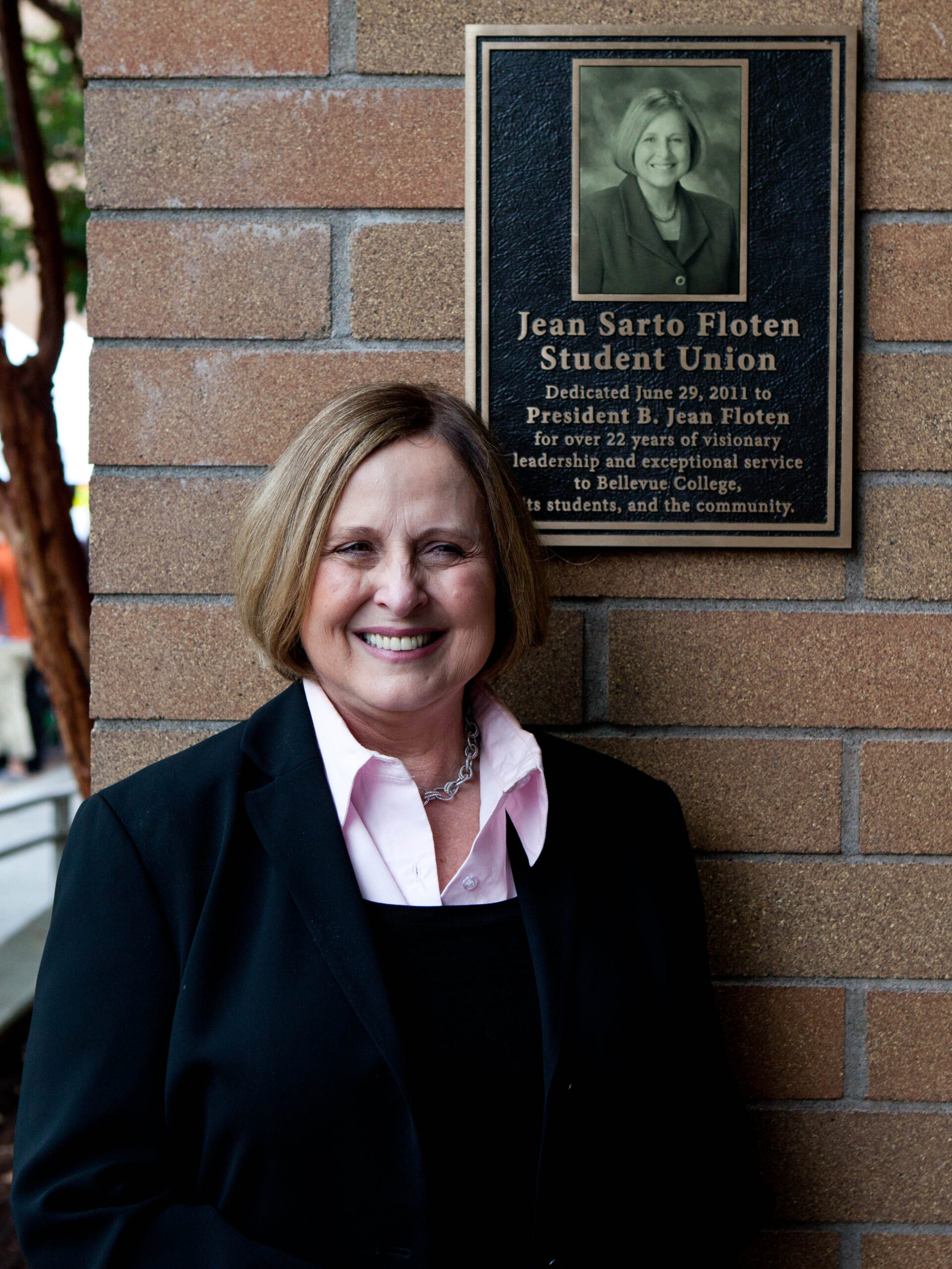 Jean Floten stands in front of brick building with plaque dedicated to her.