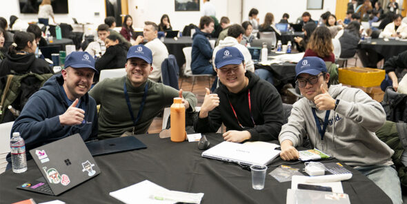 Four people sit at a table wearing Bellevue College caps, smiling and three are giving a thumbs up.