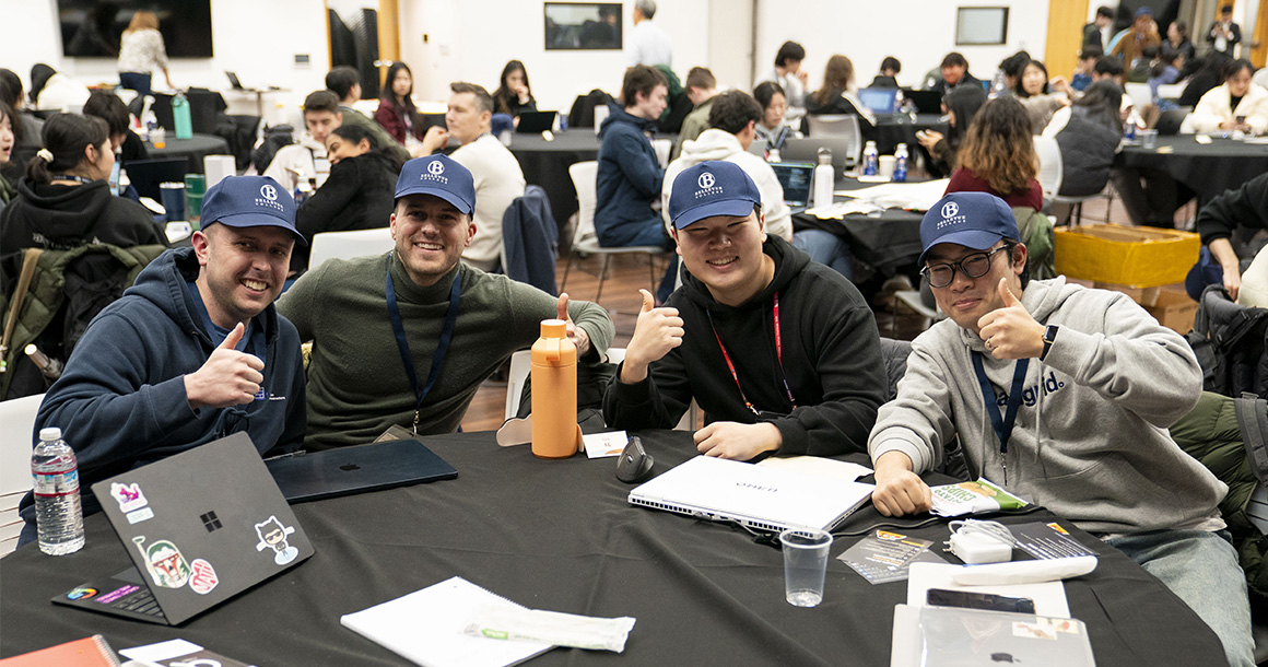 Four people sit at a table wearing Bellevue College caps, smiling and three are giving a thumbs up.