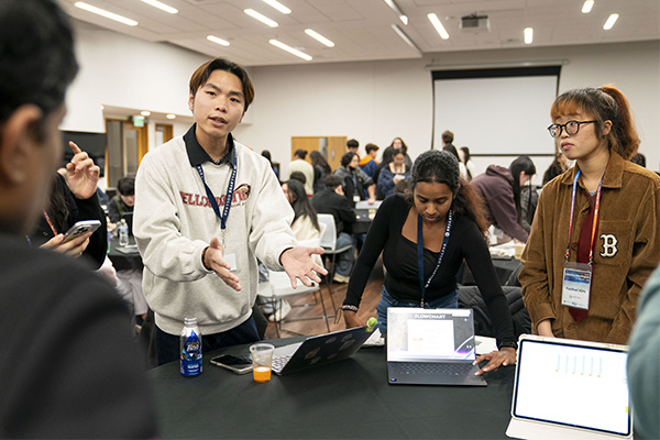 A group of people stands around a table with one person speaking and gesturing with his hands while others look on.