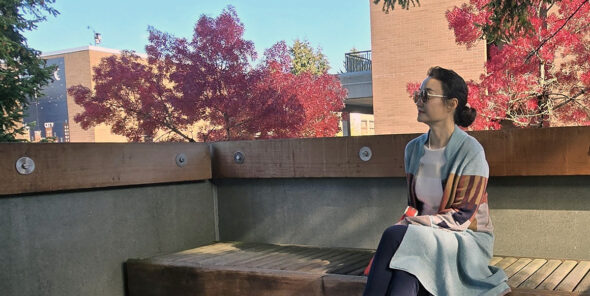 Li Liu sits on bench outside with red leaf trees behind her.