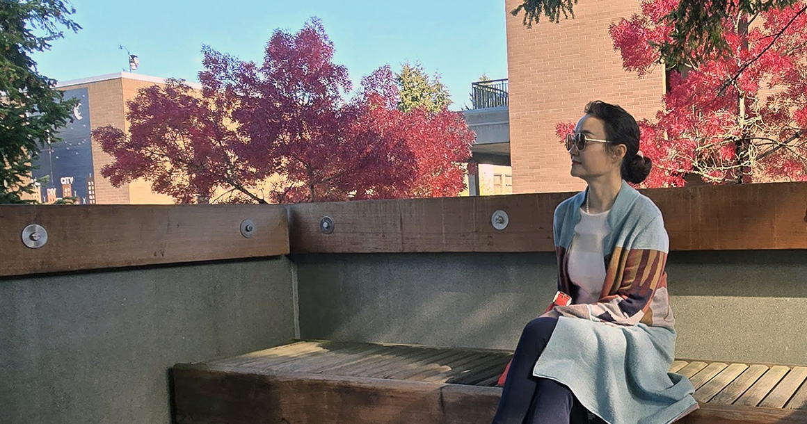Li Liu sits on bench outside with red leaf trees behind her.