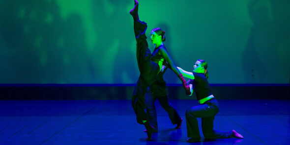 Two dancers dance on stage. One has her leg up and another is crouching beside her.