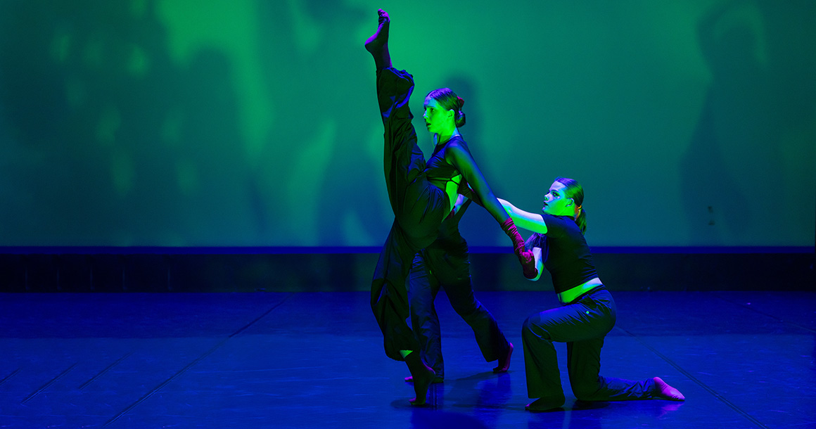 Two dancers dance on stage. One has her leg up and another is crouching beside her.