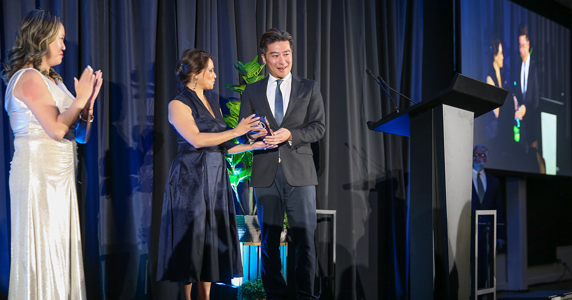 One person on stage accepts an award from another as a third person claps.