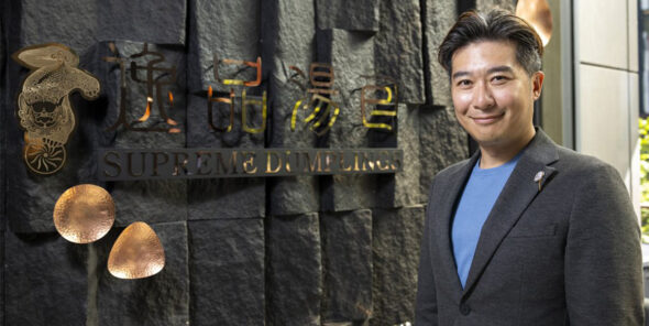 Brandon Ting stands next to Supreme Dumplings restaurant building and sign.