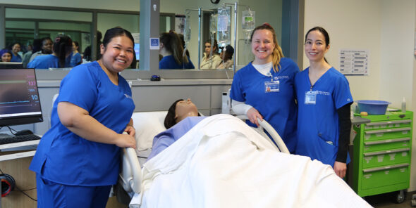 Three nursing students stand with a mannequin laying in a college lab hospital bed.