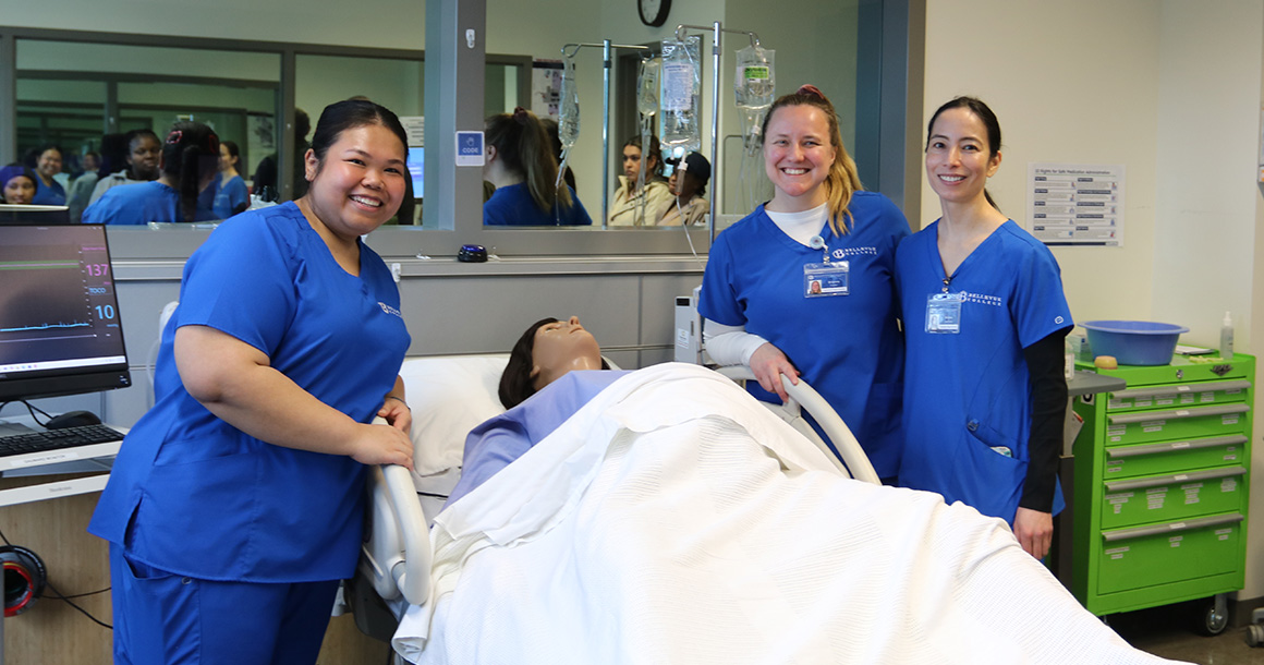 Three nursing students stand with a mannequin laying in a college lab hospital bed.