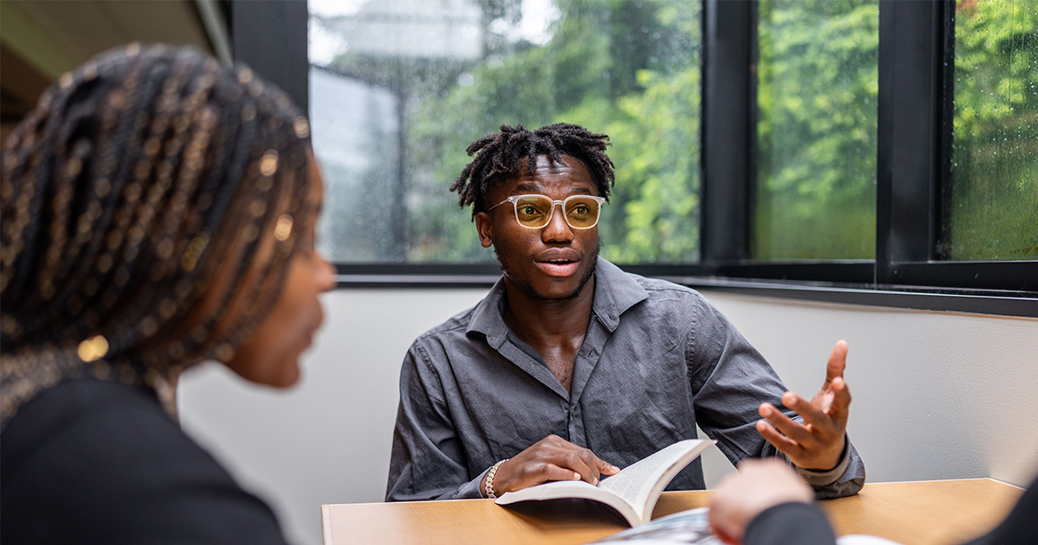 Two students sit at the table. One has a book open.