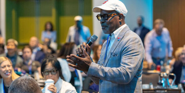 Person wearing a suit, baseball cap, and glasses speaks into a microphone in front of a sitting crowd.