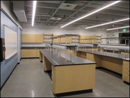 Room B113 is illuminated by four long, narrow tube lights. A white board is featured prominently at the front of the room, and several workstations are spaced out in the center. Along a far wall, there are mounted cabinets and additional countertops with storage below.