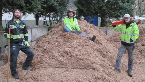 Two members of the BC Grounds Crew pose with tools and safety vests by a large pile of mulch. A third crew member sits atop the pile.