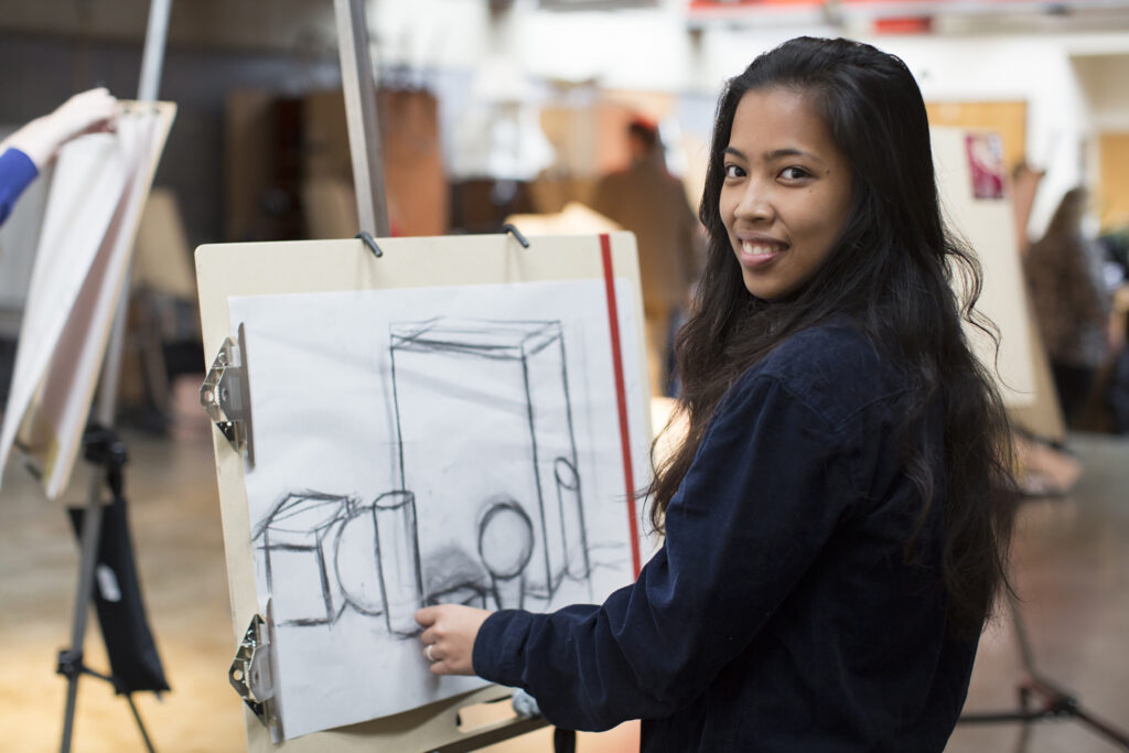 A student displays a drawing at her easel.