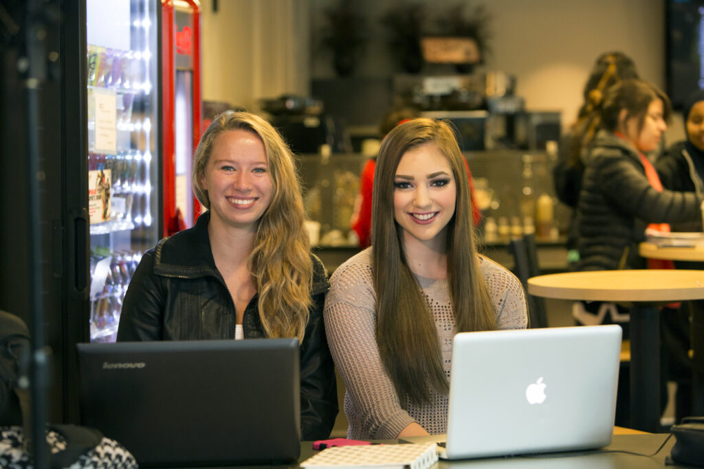 Two young women smile in the cafe while working on computers.