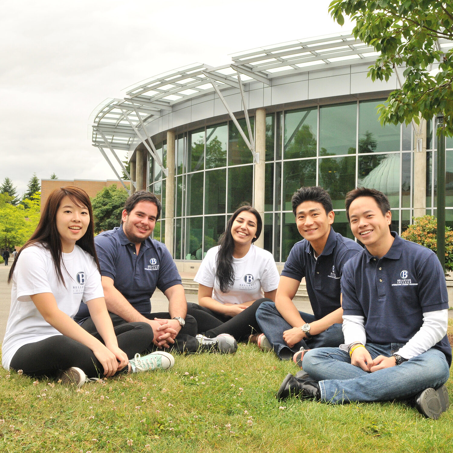 A group of five students sit close together in a grassy space in front of a campus building.