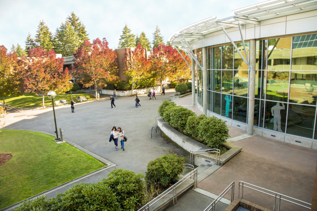 The exterior of Bellevue College's Student Center is seen from a vantage point a few stories high from a neighboring building. About 10 people are seen walking through the courtyard and lounging on benches. Around them are several grassy spots, shrubs, and trees with fall foliage.