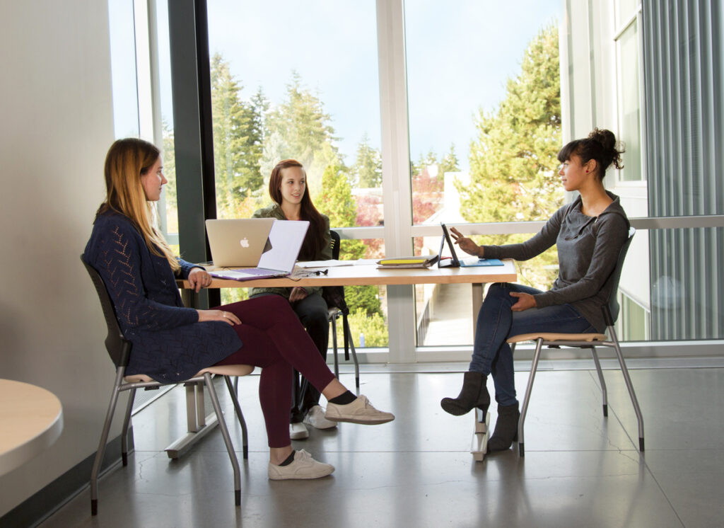 Three students face each other while sitting together at a table within a campus building. They have their laptops open and appear to be engaged in conversation. There is a view of campus through the large window behind them.