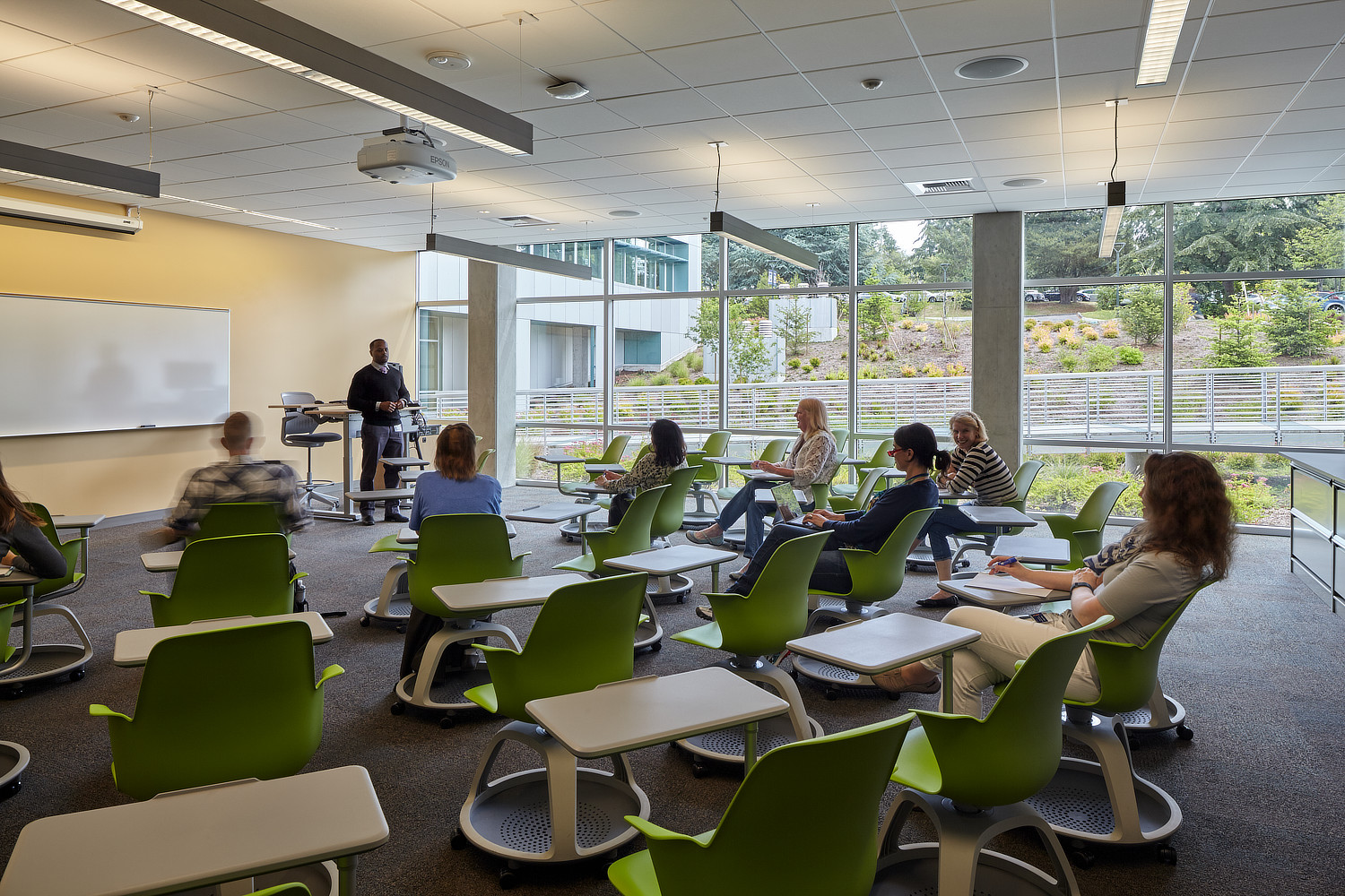 Eight students sit at desks in classroom facing the instructor, who is standing at a podium in the front of the room.