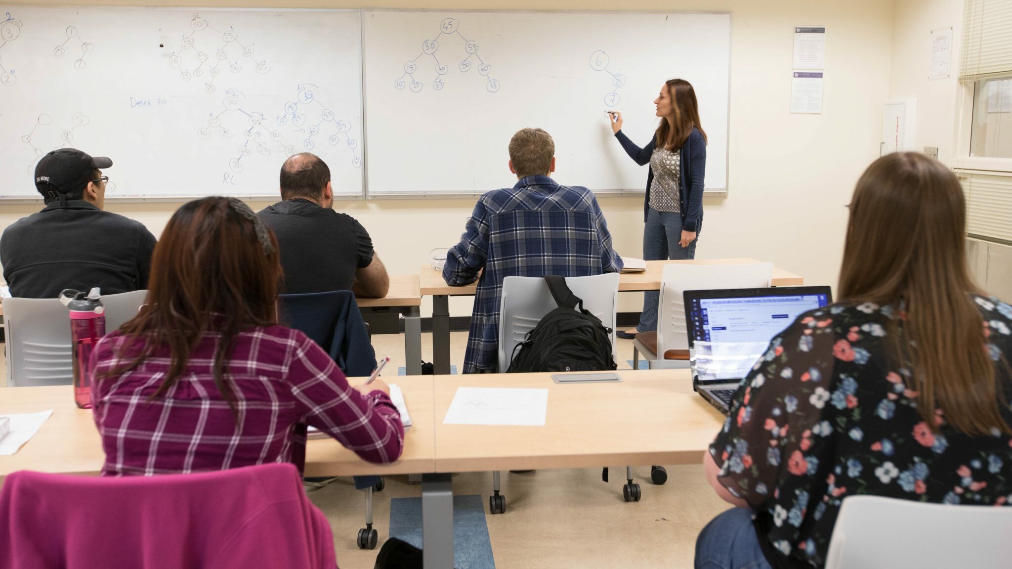 Five students sit at desks listening to the professor, who writes on the whiteboard