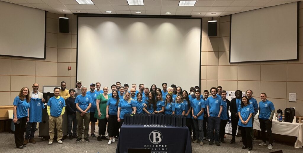 Over 40 Bellevue College Computer Science students and faculty members stand together in a large classroom for the 2023 senior capstone competition. Many people are wearing a blue t-shirt featuring the Bellevue College Computer Science logo. The group smiles together toward the camera.