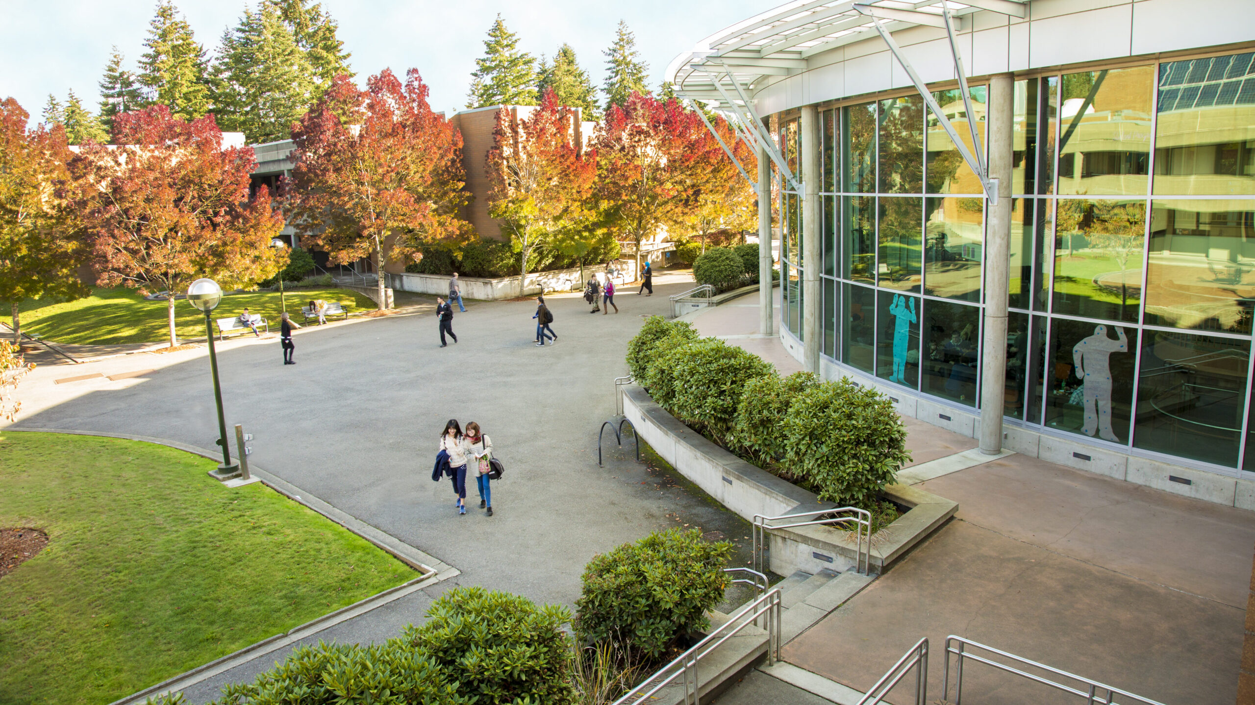 The exterior of Bellevue College's Student Center is seen from a vantage point a few stories high from a neighboring building. About 10 people are seen walking through the courtyard and lounging on benches. Around them are several grassy spots, shrubs, and trees with fall foliage.
