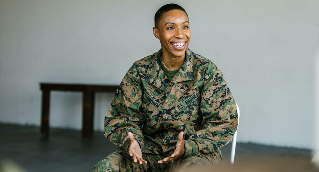 A smiling US Marine sits in a chair and gestures in conversation.