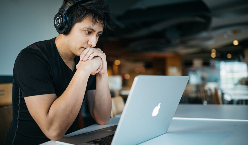 A student wears headphones while looking thoughtfully at a laptop.
