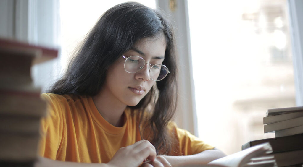 A student sits by a window and studies while surrounded by stacks of books.