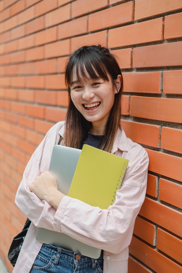 Student holding class books leaning against a brick wall