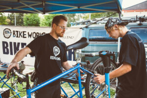 Two Bike Works Volunteers are working on a bike that is on a bike stand. A Bike Works banner is in the background.