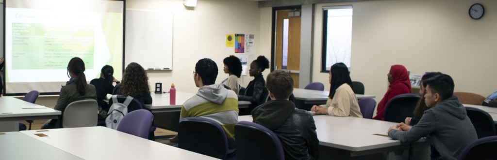 Students in a classroom are looking at a presentation on a projector screen