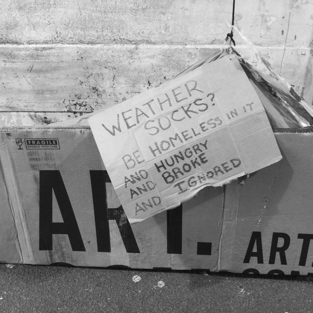 Black and white photo of a cardboard sign that says "Weather Sucks? Be homeless in it and hungry and broke and ignored"