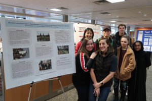 Six students and their instructor stand next to a large poster about their service-learning project for the Fall 2019 Making Learning Visible student showcase