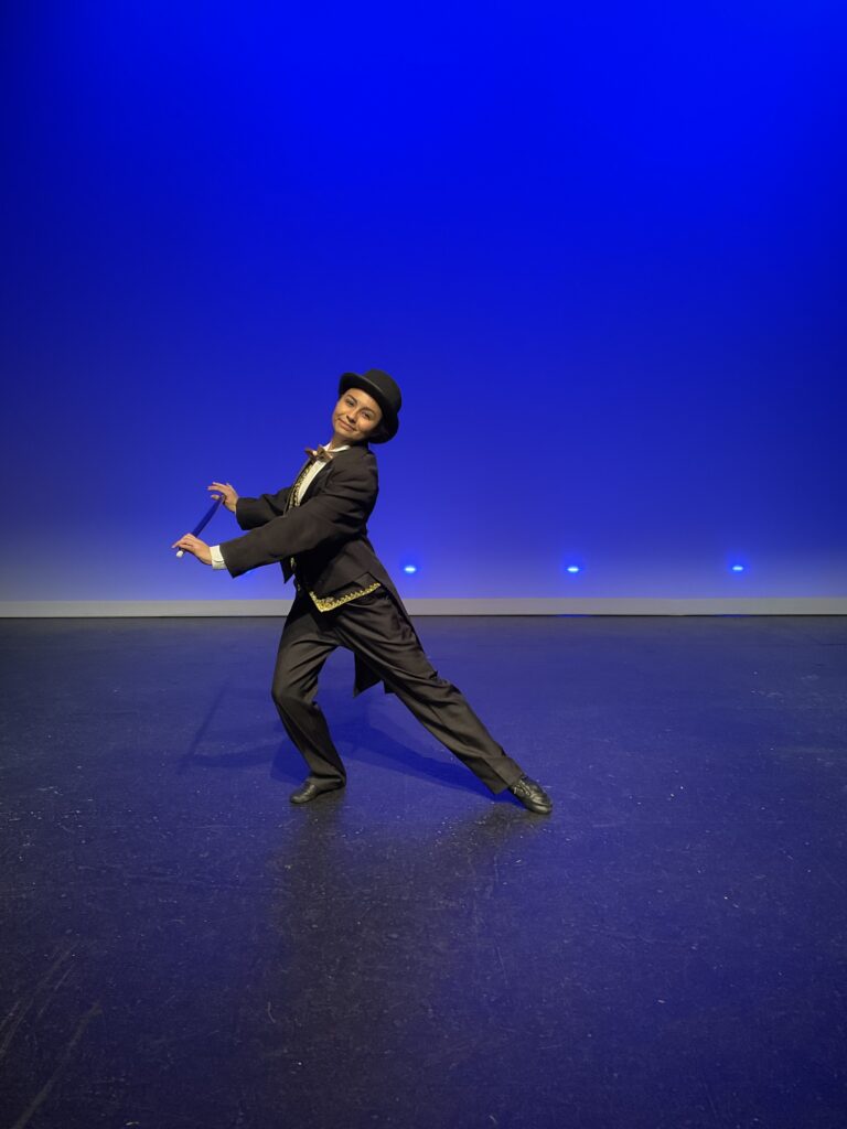 Student in black tuxedo and top hat making an artistic dance pose on a stage with blue background